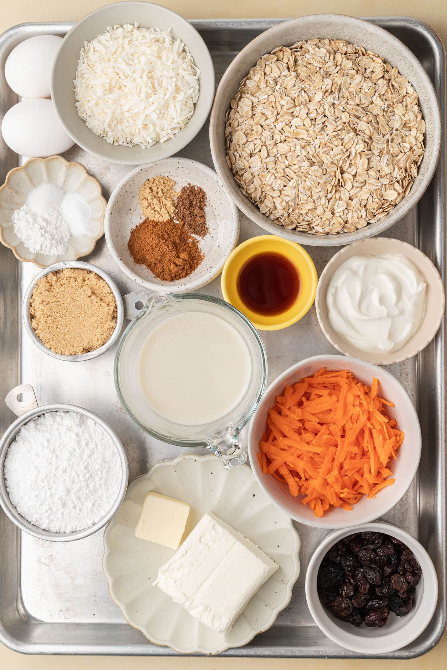 Baking ingredients on a tray, including oats, eggs, shredded carrot, spices, raisins, and various bowls of dairy.