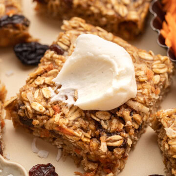 A square oat bar topped with cream, surrounded by dried fruit and oats on a light surface.