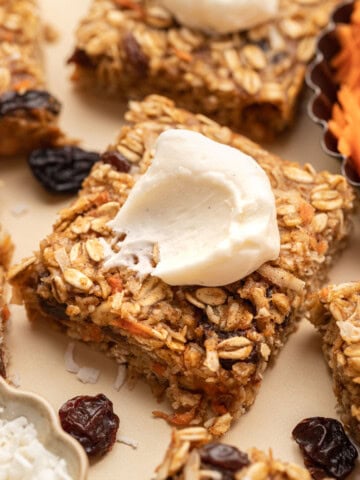 A square oat bar topped with cream, surrounded by dried fruit and oats on a light surface.