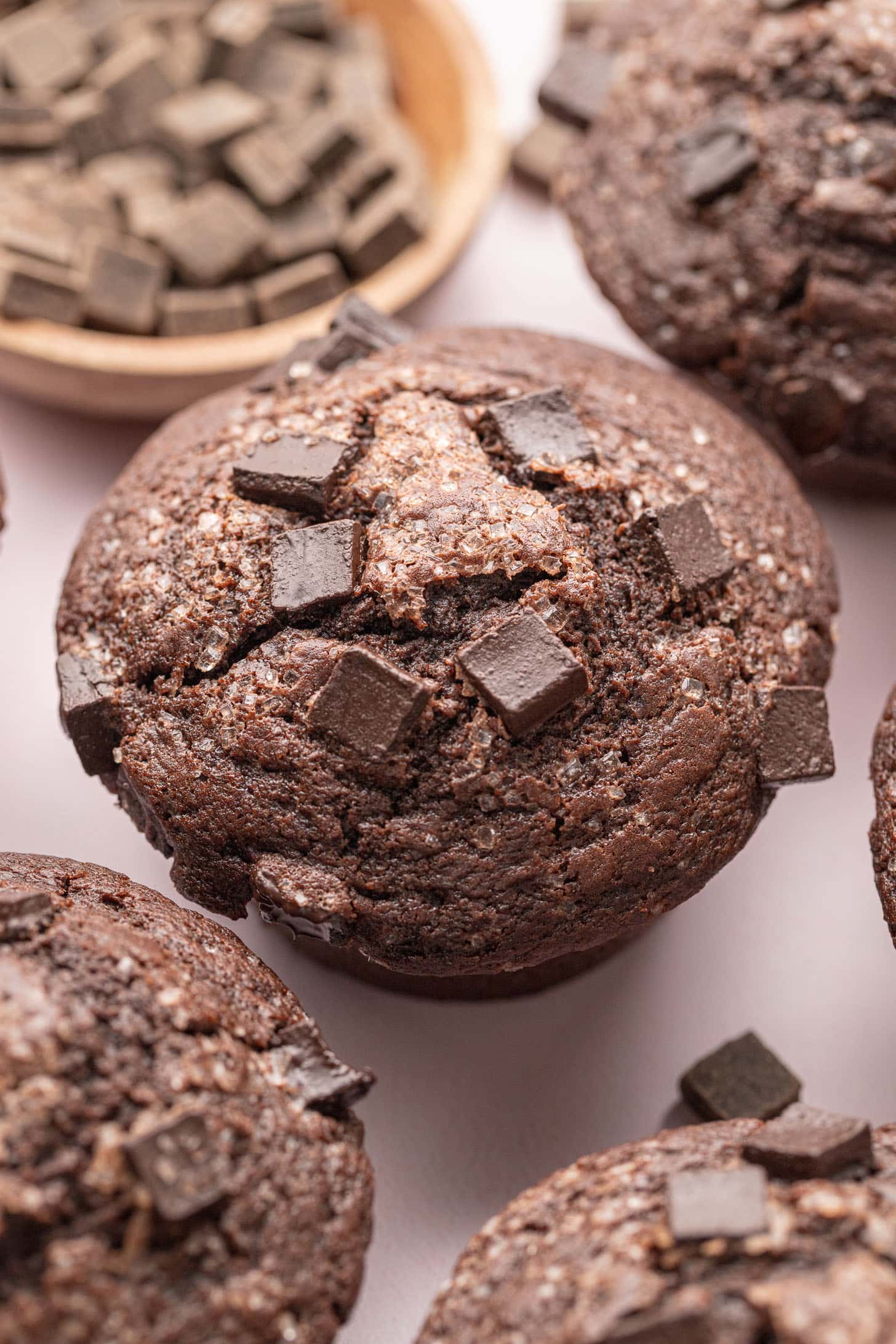 Close-up of chocolate muffins topped with chocolate chunks, with a bowl of chocolate pieces in the background.