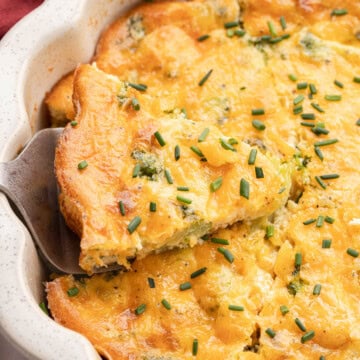 A slice of cheesy broccoli quiche is being served from a white baking dish, topped with chopped chives.