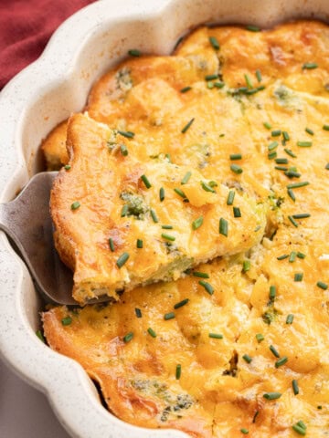 A slice of cheesy broccoli quiche is being served from a white baking dish, topped with chopped chives.