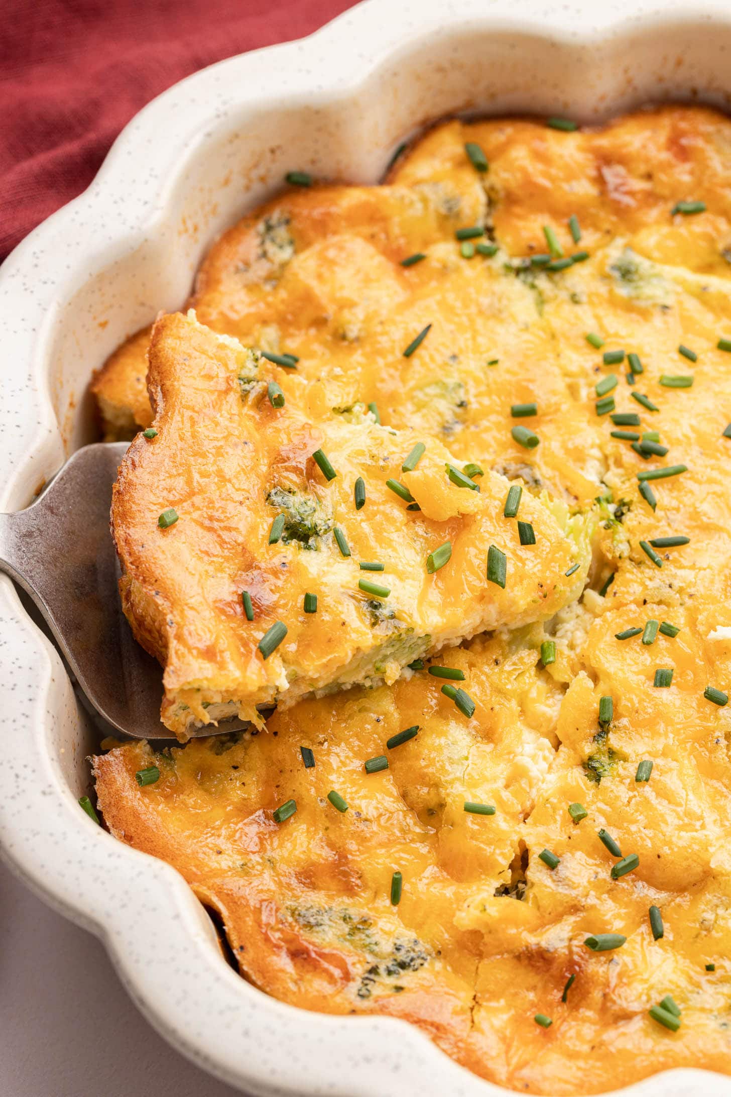 A slice of cheesy broccoli quiche is being served from a white baking dish, topped with chopped chives.