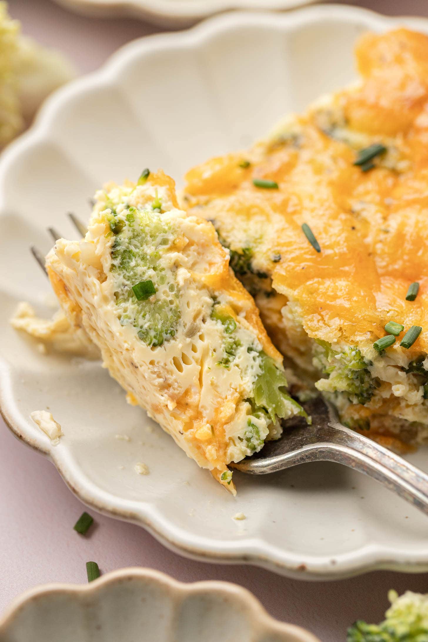 A fork holds a cheesy broccoli and egg casserole slice on a white scalloped plate, topped with chives.