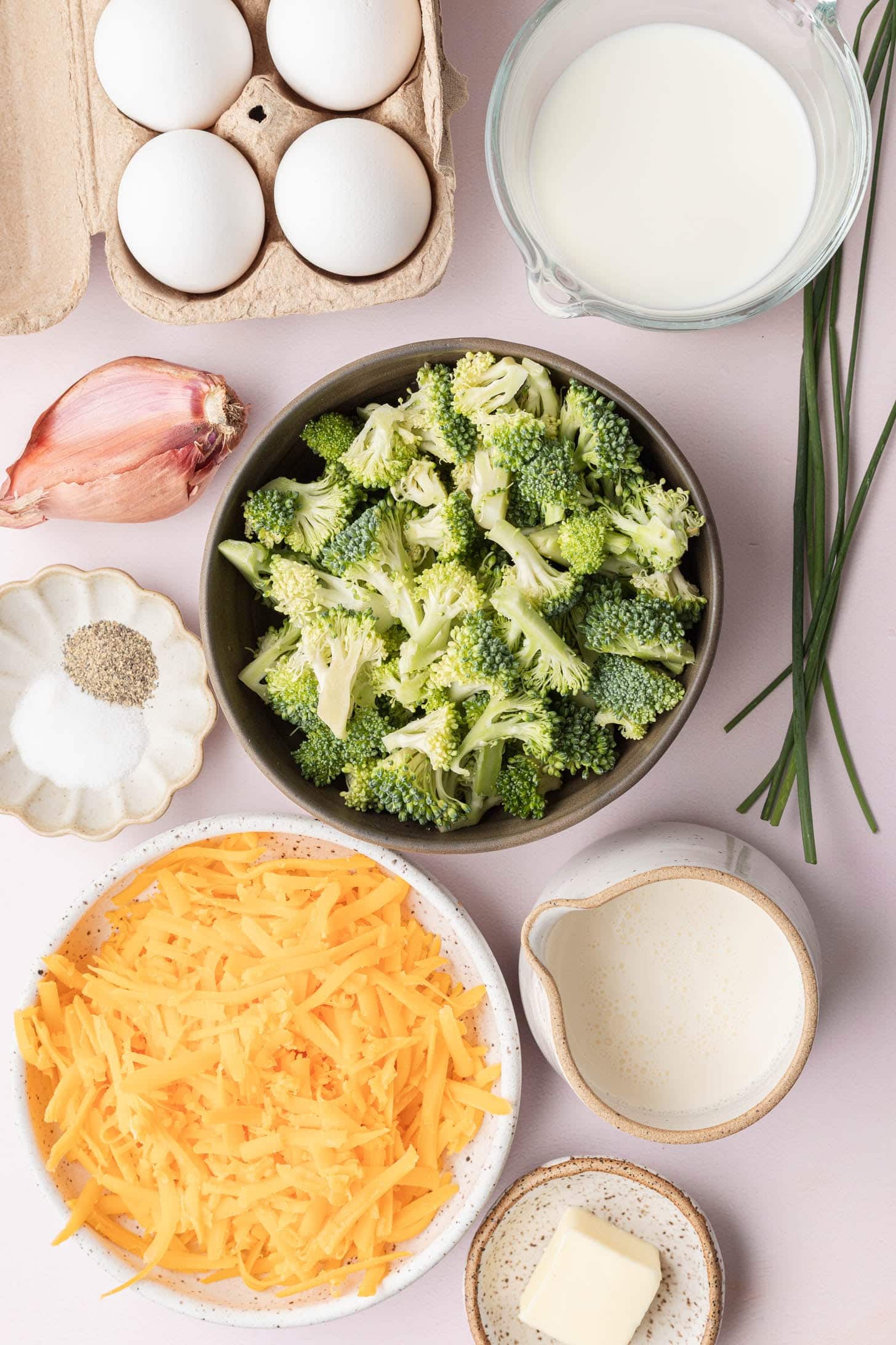 Eggs, broccoli, milk, cheddar, shallot, chives, butter, cream, salt, and pepper arranged on a table.