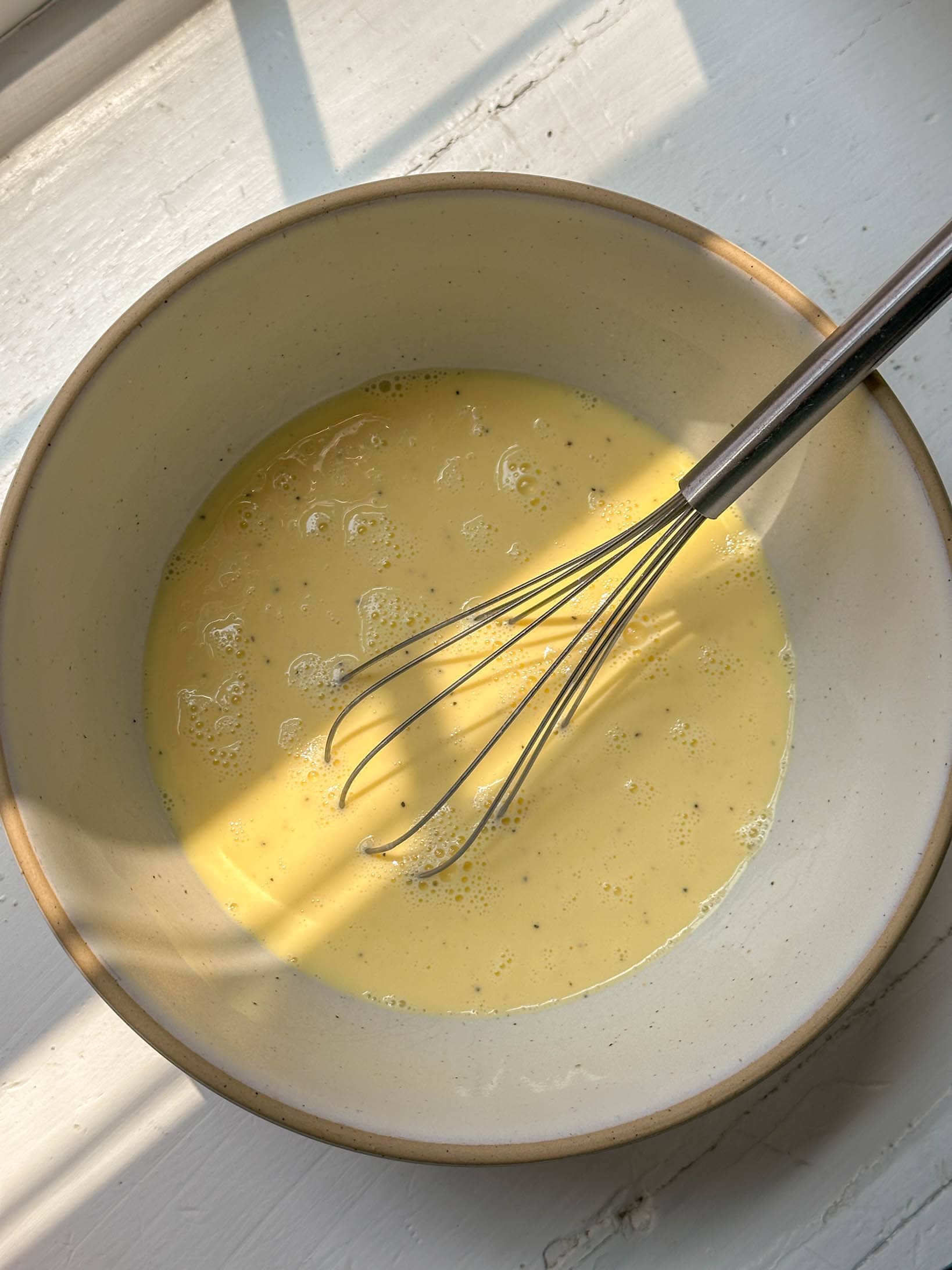 A metal whisk in a bowl of beaten eggs on a sunlit surface.