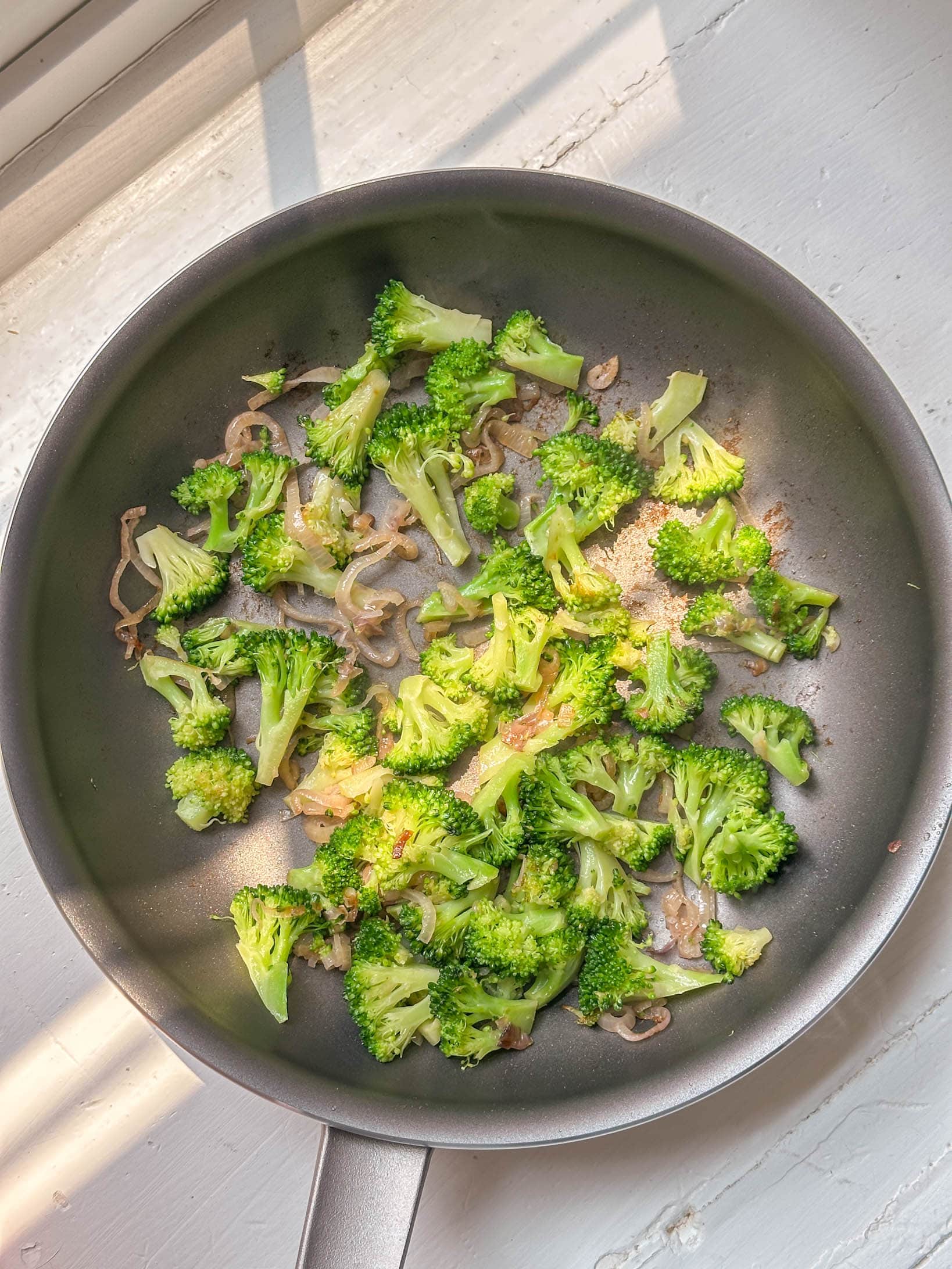 Broccoli and sliced onions sautéing in a nonstick pan on a sunlit white countertop.