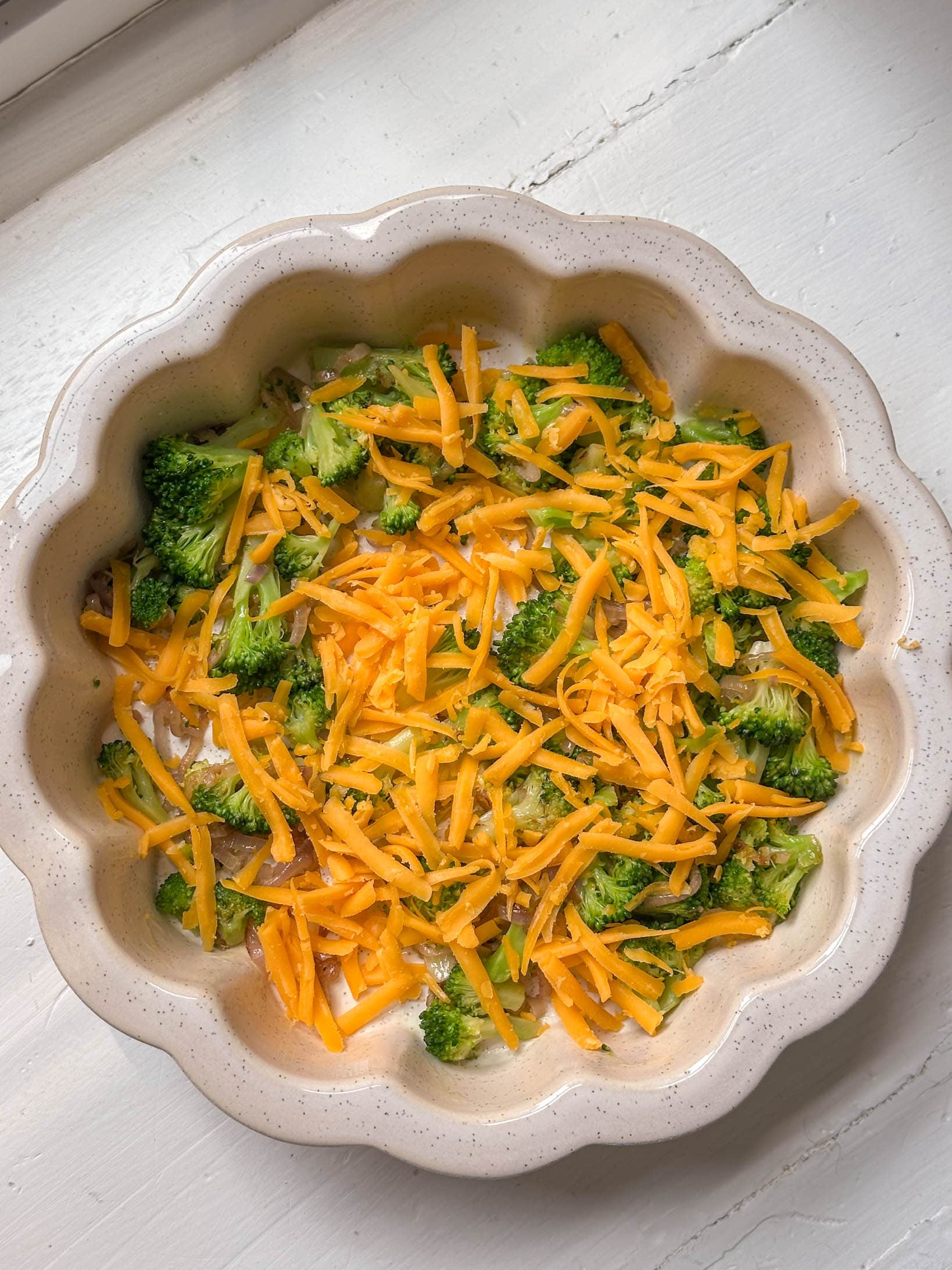A round baking dish filled with chopped broccoli and shredded cheddar cheese on a white surface.