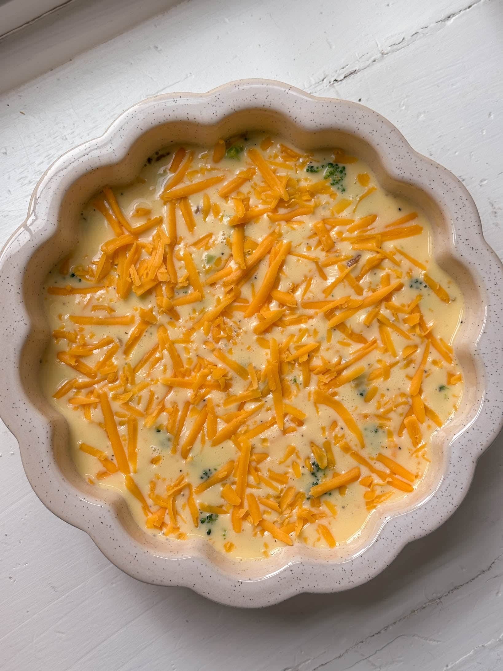 Unbaked quiche in a scalloped dish, topped with shredded cheddar cheese and visible broccoli pieces.