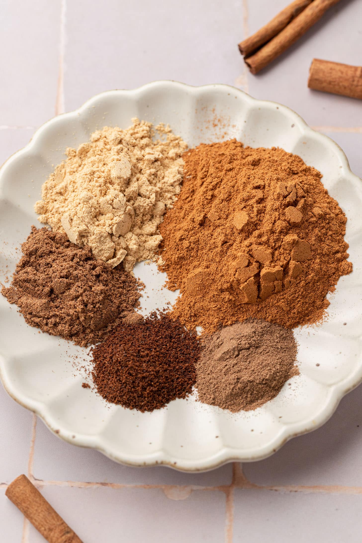A white plate holds six different ground spices, with cinnamon sticks placed beside the plate.