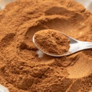 A spoon scoops ground cinnamon from a white scalloped bowl filled with the spice.