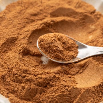 A spoon scoops ground cinnamon from a white scalloped bowl filled with the spice.