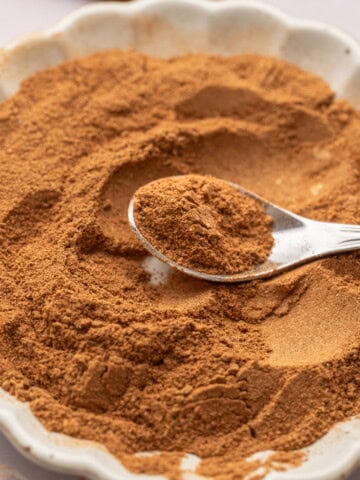 A spoon scoops ground cinnamon from a white scalloped bowl filled with the spice.