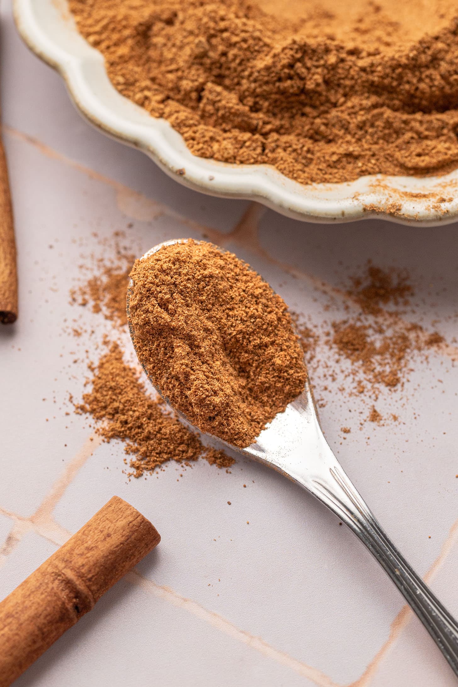 A spoonful of ground cinnamon on a table next to cinnamon sticks and a plate of more cinnamon.