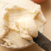 A knife spreads creamy butter in a bowl, with a honey dipper and a small dish in the background.