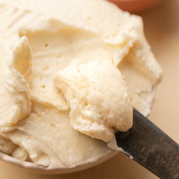 A knife spreads creamy butter in a bowl, with a honey dipper and a small dish in the background.