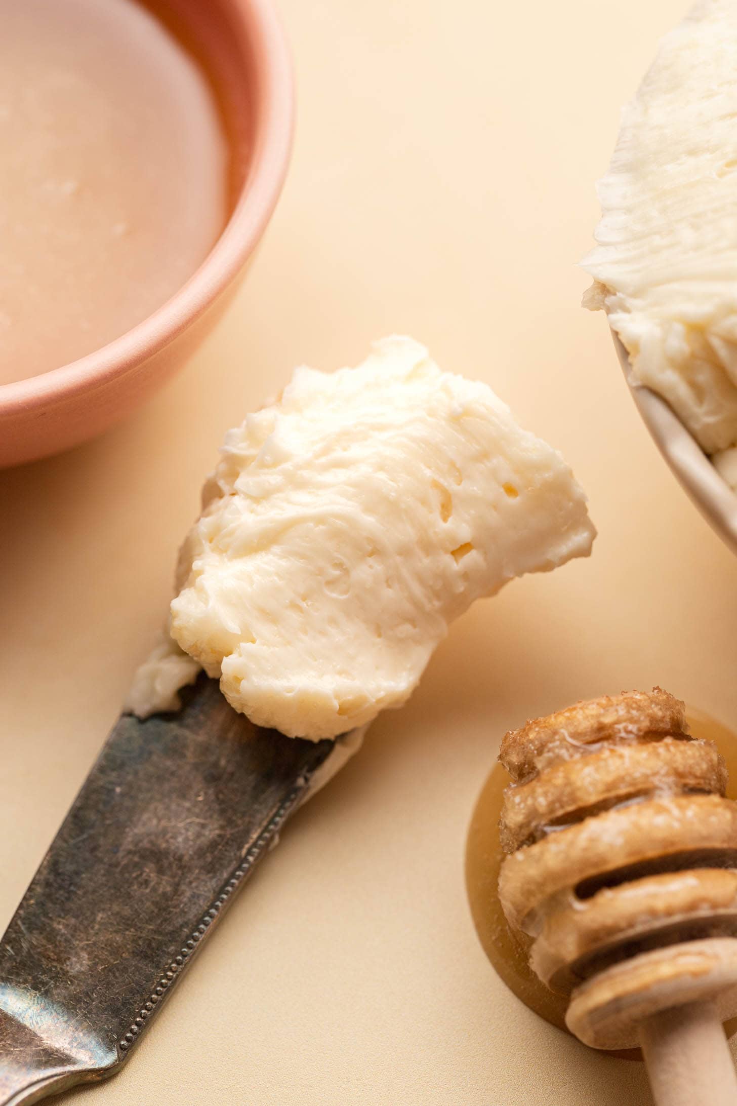 Close-up of a butter knife with whipped butter, a honey dipper, and a bowl of honey on a beige surface.