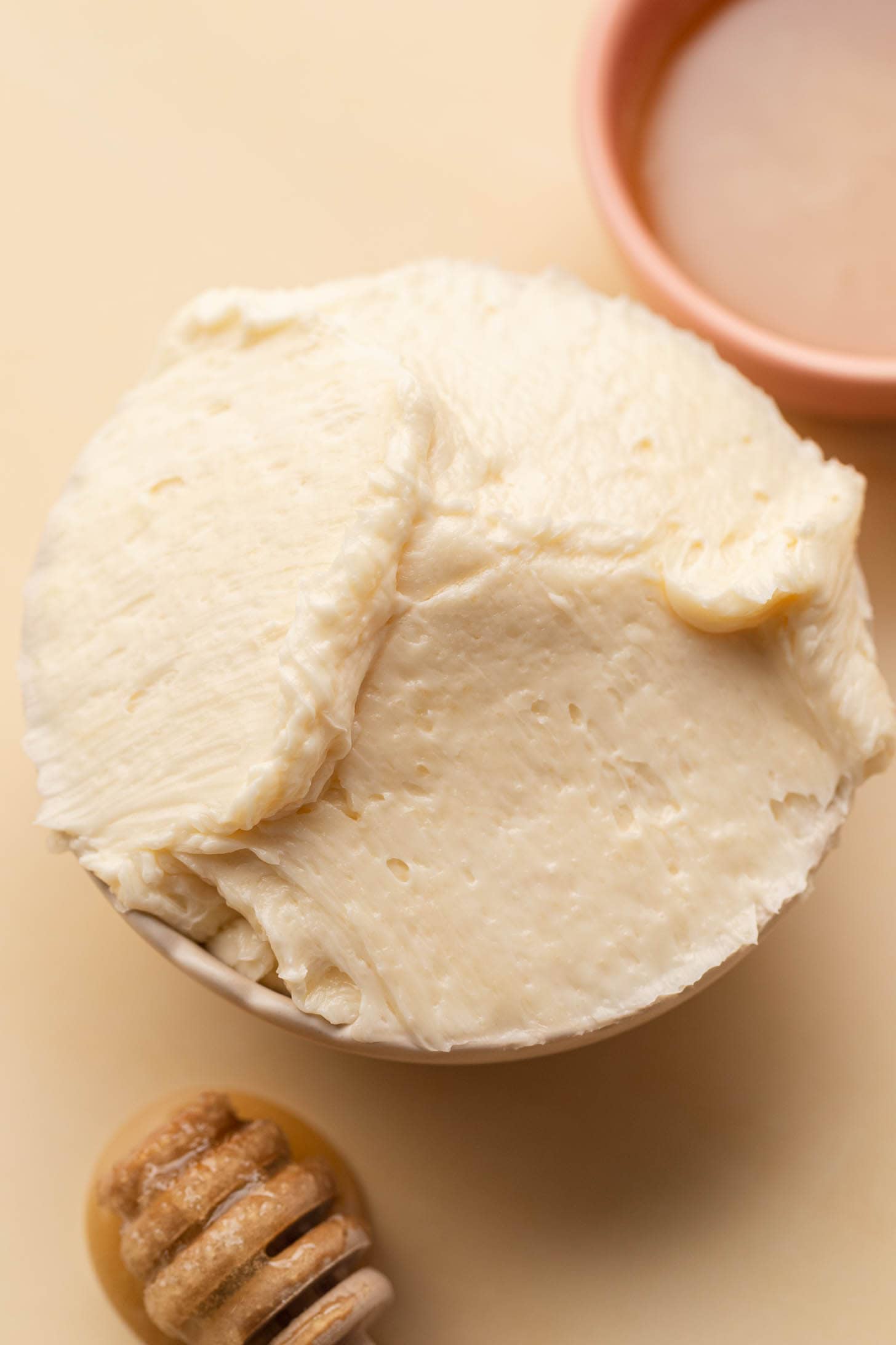 A bowl of whipped butter with a honey dipper and a small dish of honey nearby on a beige surface.