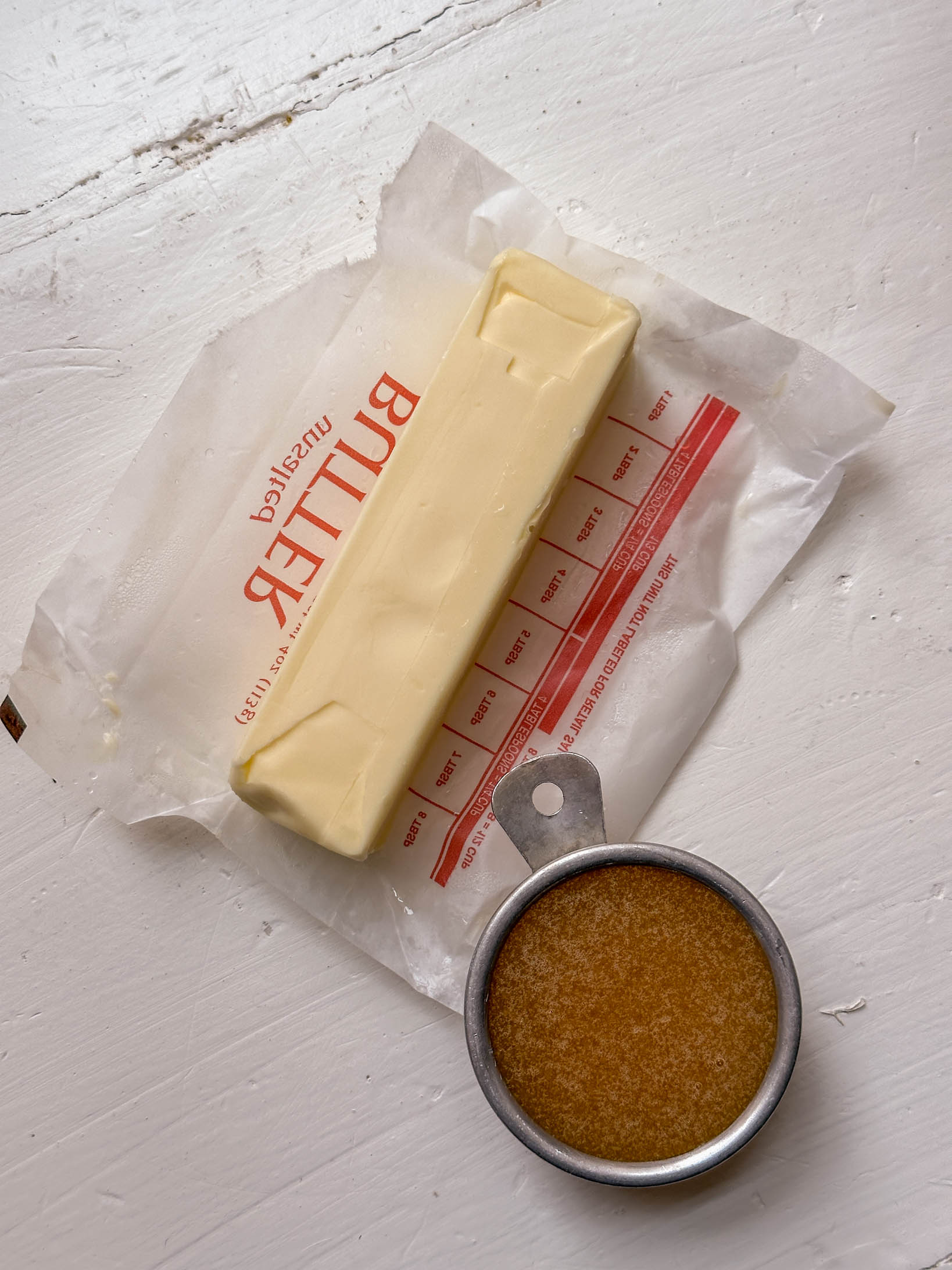 A stick of butter on its wrapper next to a metal measuring cup filled with melted butter.