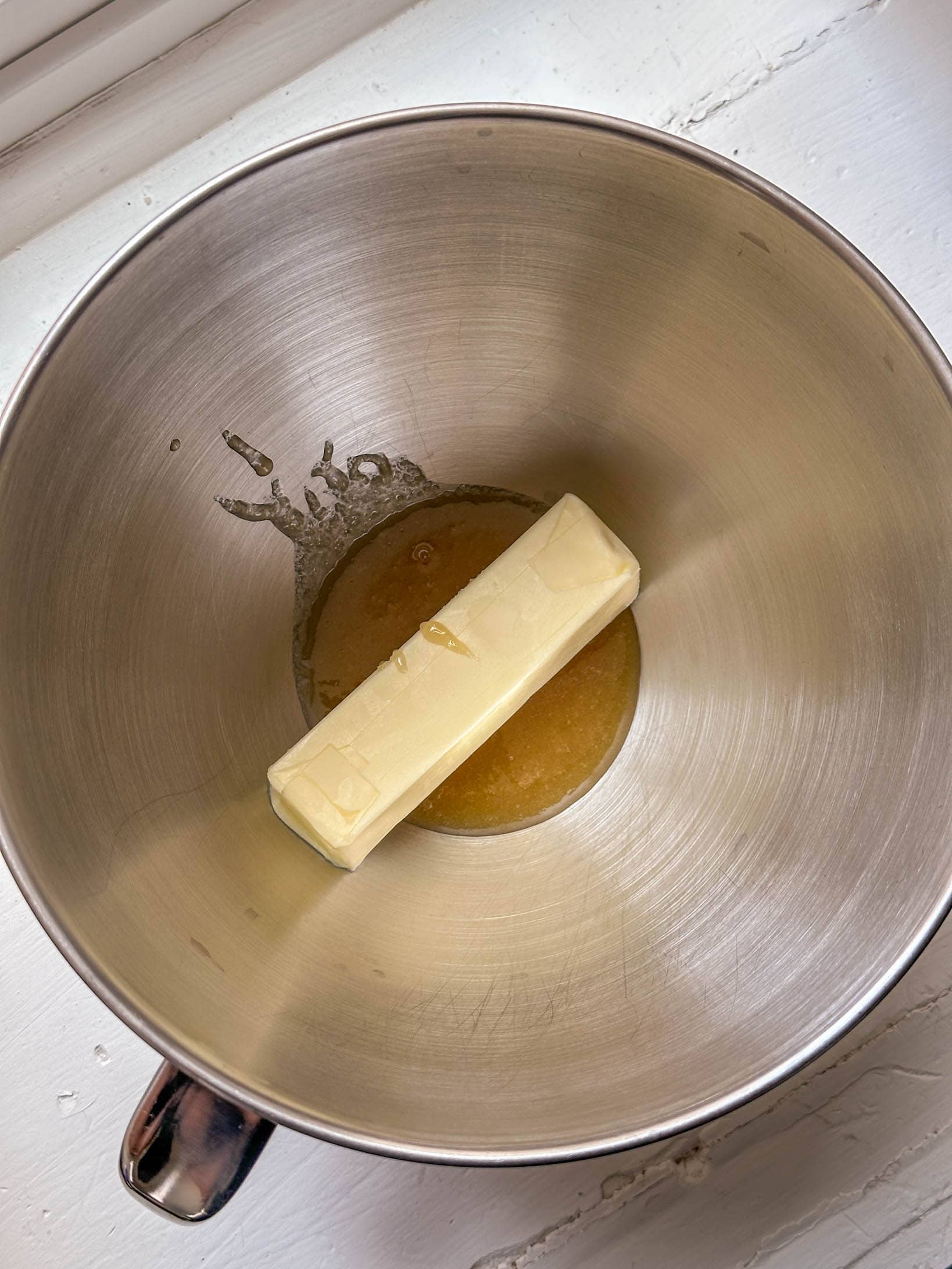A stick of butter and melted brown sugar in a metal mixing bowl on a white countertop.