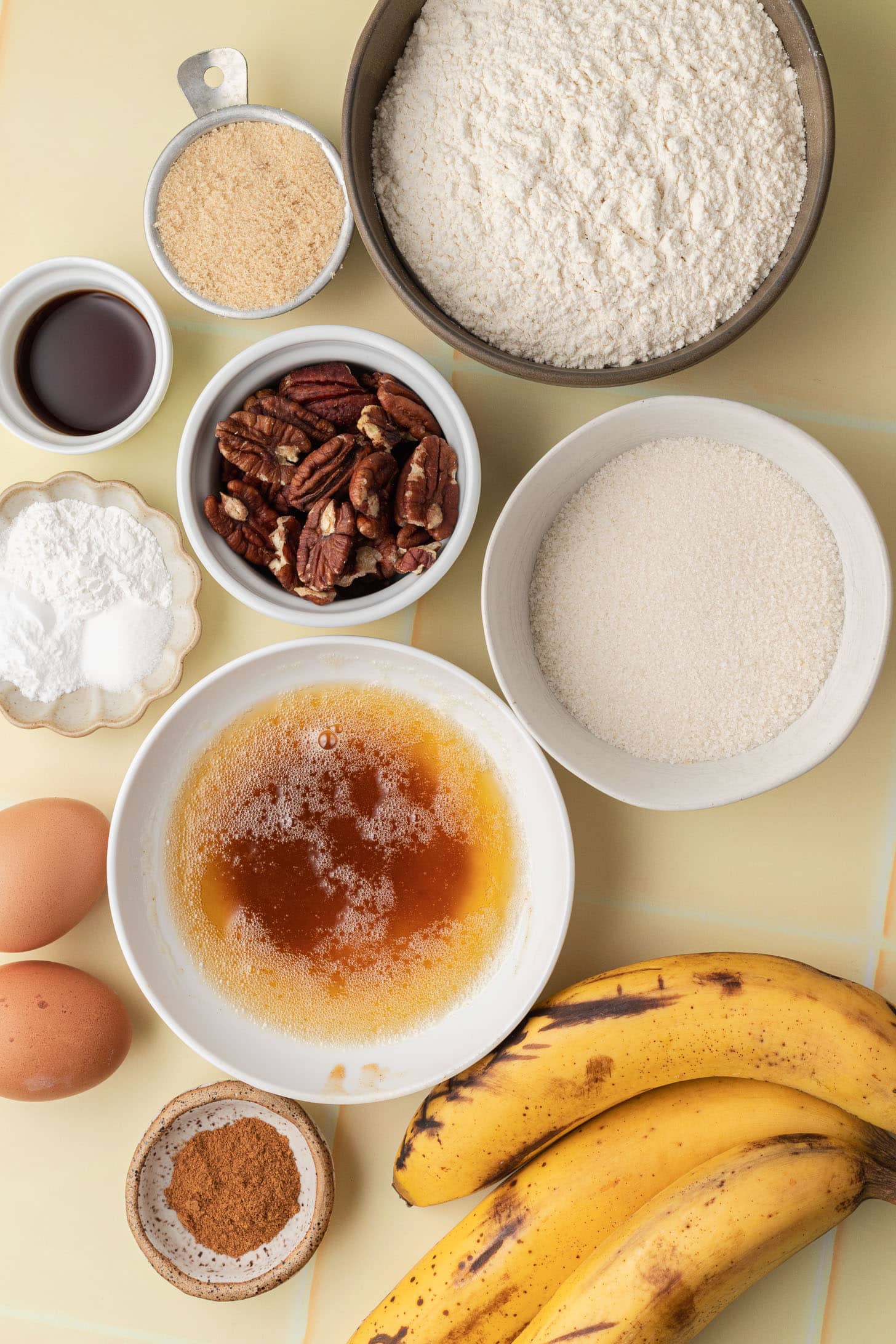 Bowls with flour, sugar, pecans, eggs, bananas, spices, vanilla, and melted butter on a light surface.