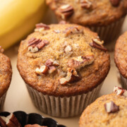 Banana nut muffins topped with chopped pecans, with bananas and a small dish of pecans in the background.