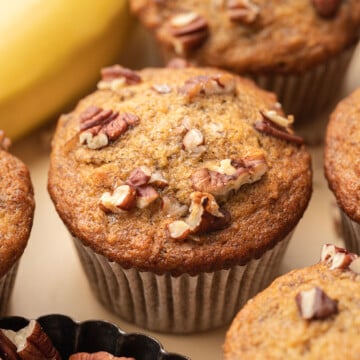 Banana nut muffins topped with chopped pecans, with bananas and a small dish of pecans in the background.