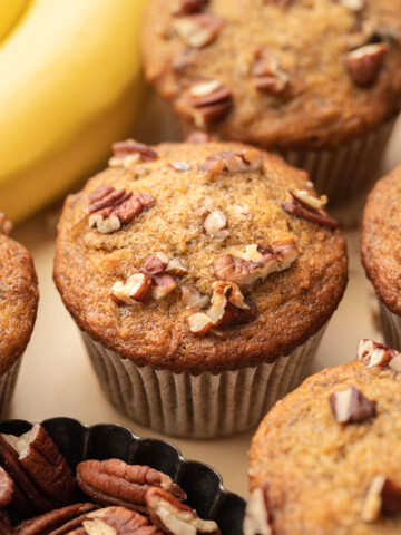 Banana nut muffins topped with chopped pecans, with bananas and a small dish of pecans in the background.
