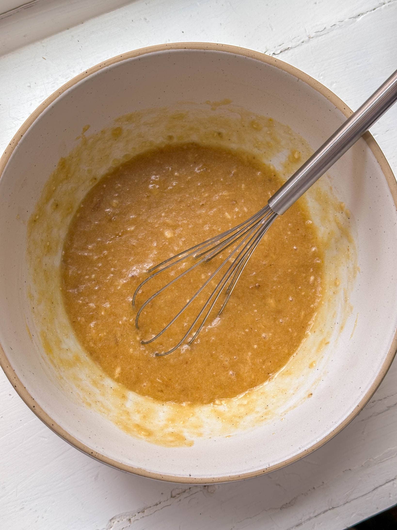 A whisk in a bowl of light brown batter sits on a white surface.