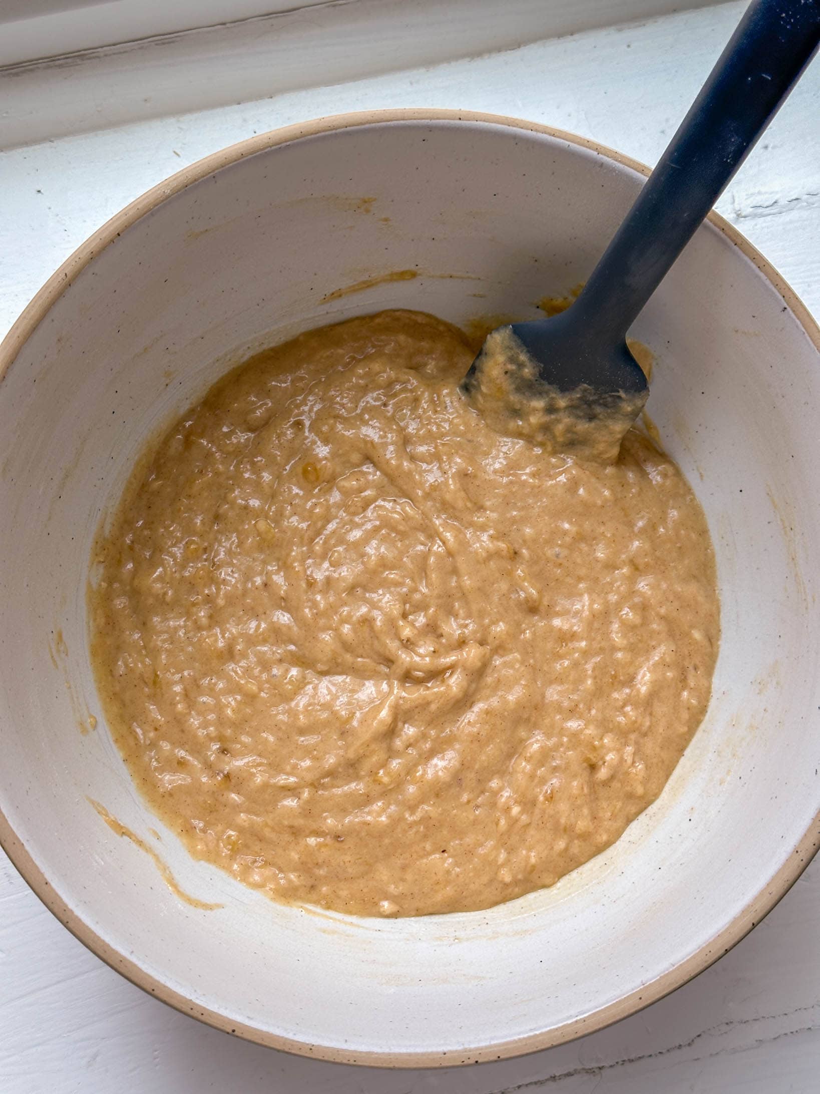 A white bowl with tan batter and a dark blue spatula resting inside, on a white surface.