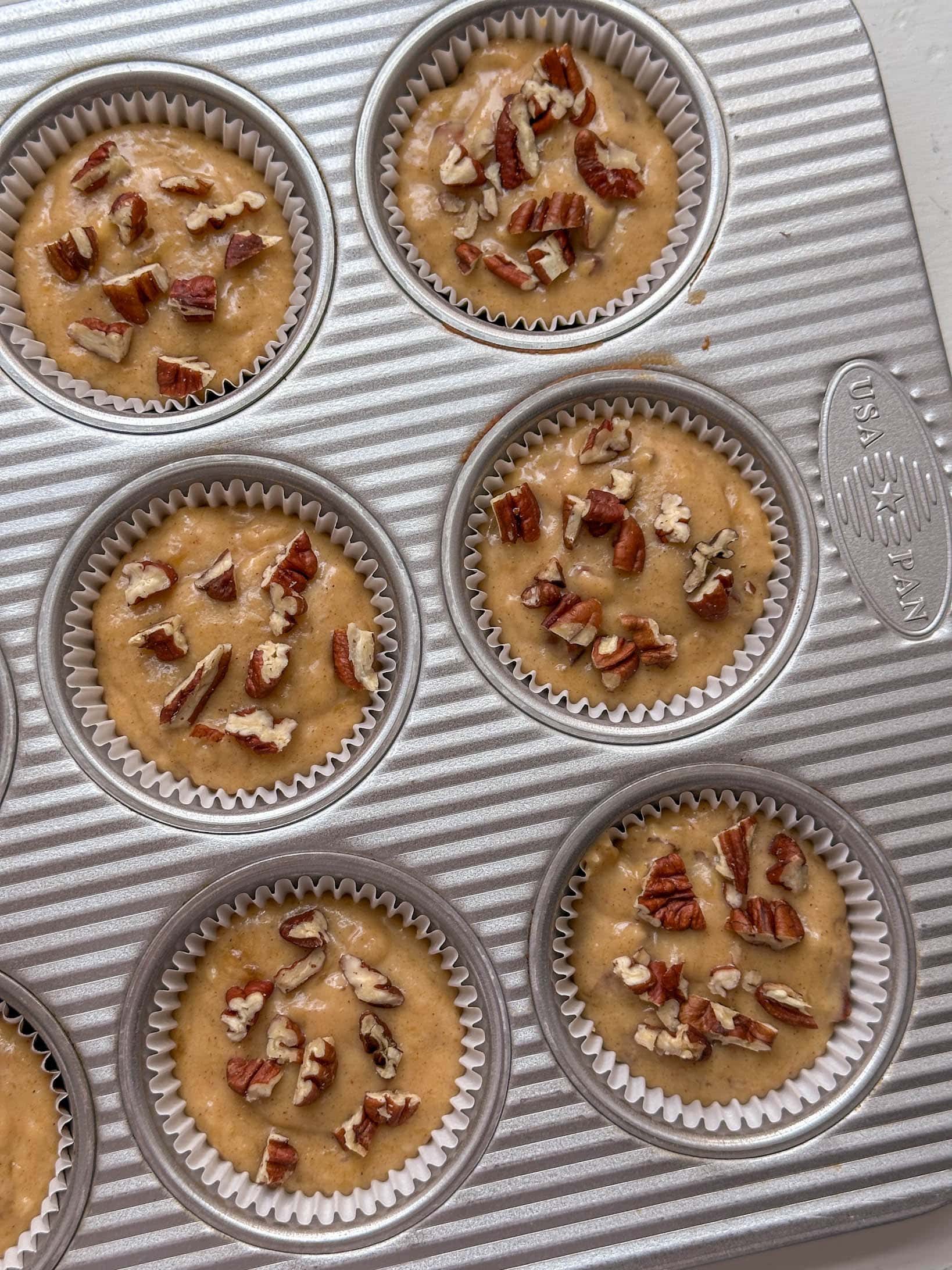 Muffin tin with unbaked batter in liners, topped with chopped pecans, ready to bake.