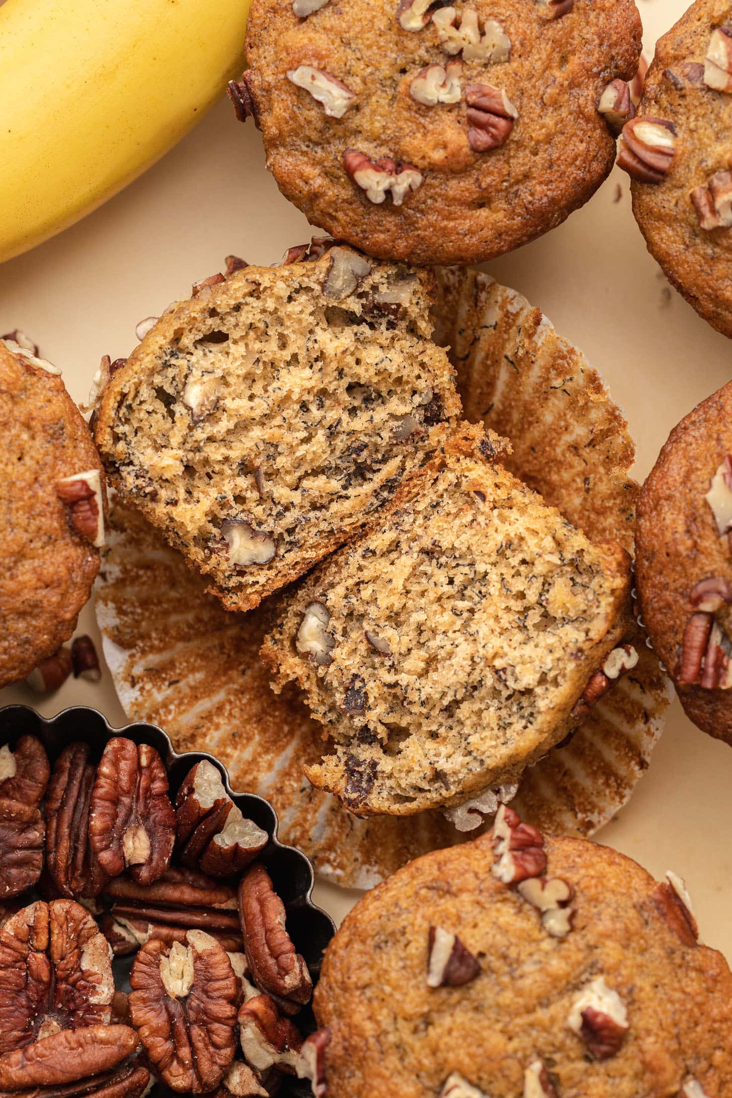 Banana nut muffin cut in half on a wrapper, surrounded by whole muffins, pecans, and a banana.