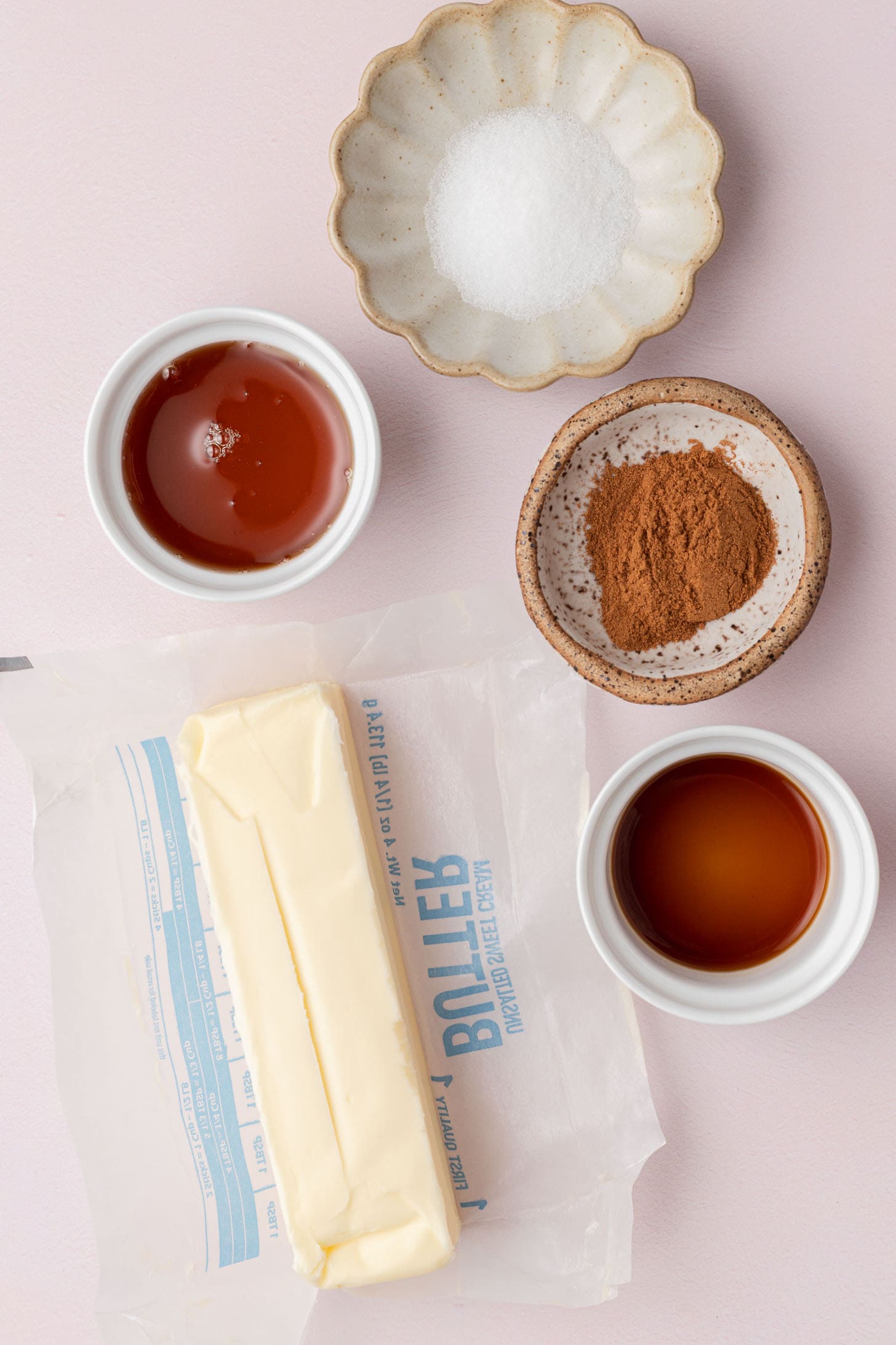 A stick of butter, cinnamon, vanilla, honey, and sugar in small bowls on a pink surface.