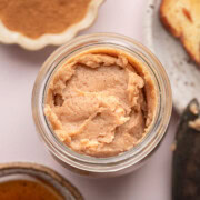 Jar of cinnamon butter, with honey, ground cinnamon, and a slice of bread nearby on a light surface.