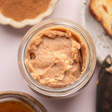 Jar of cinnamon butter, with honey, ground cinnamon, and a slice of bread nearby on a light surface.