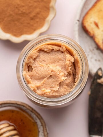 Jar of cinnamon butter, with honey, ground cinnamon, and a slice of bread nearby on a light surface.