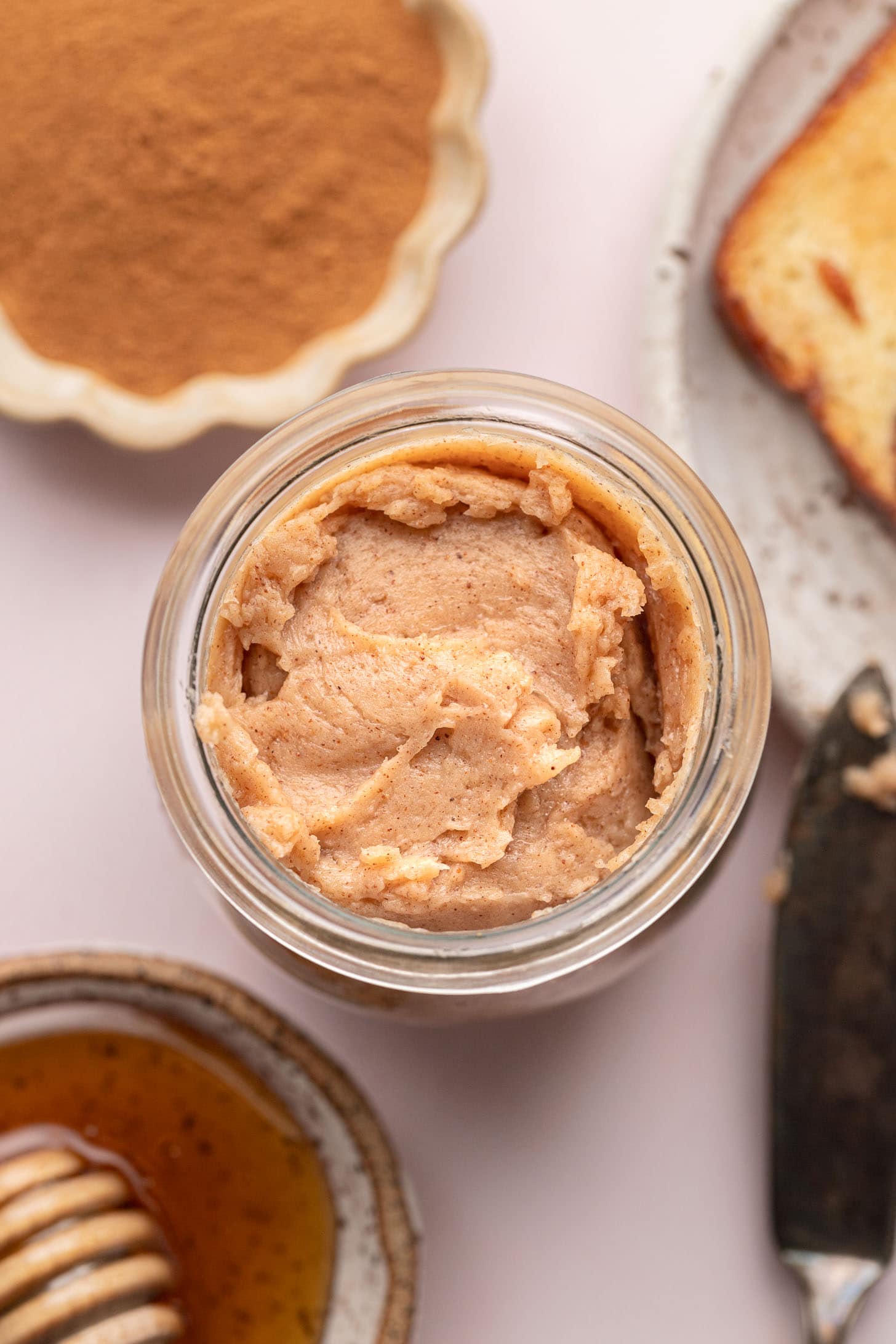 Jar of cinnamon butter, with honey, ground cinnamon, and a slice of bread nearby on a light surface.