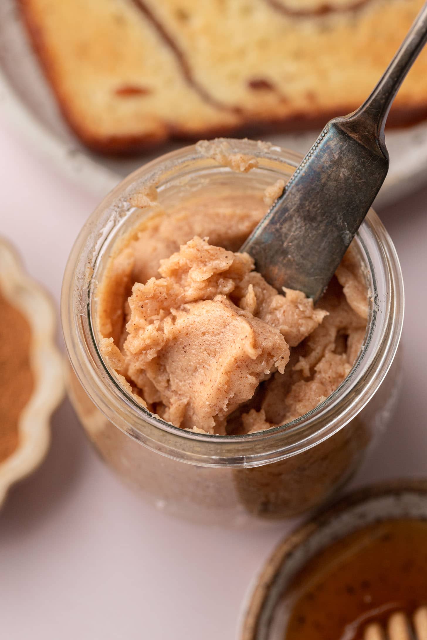 A spoon scoops cinnamon butter from a jar, with bread and spices in the background.
