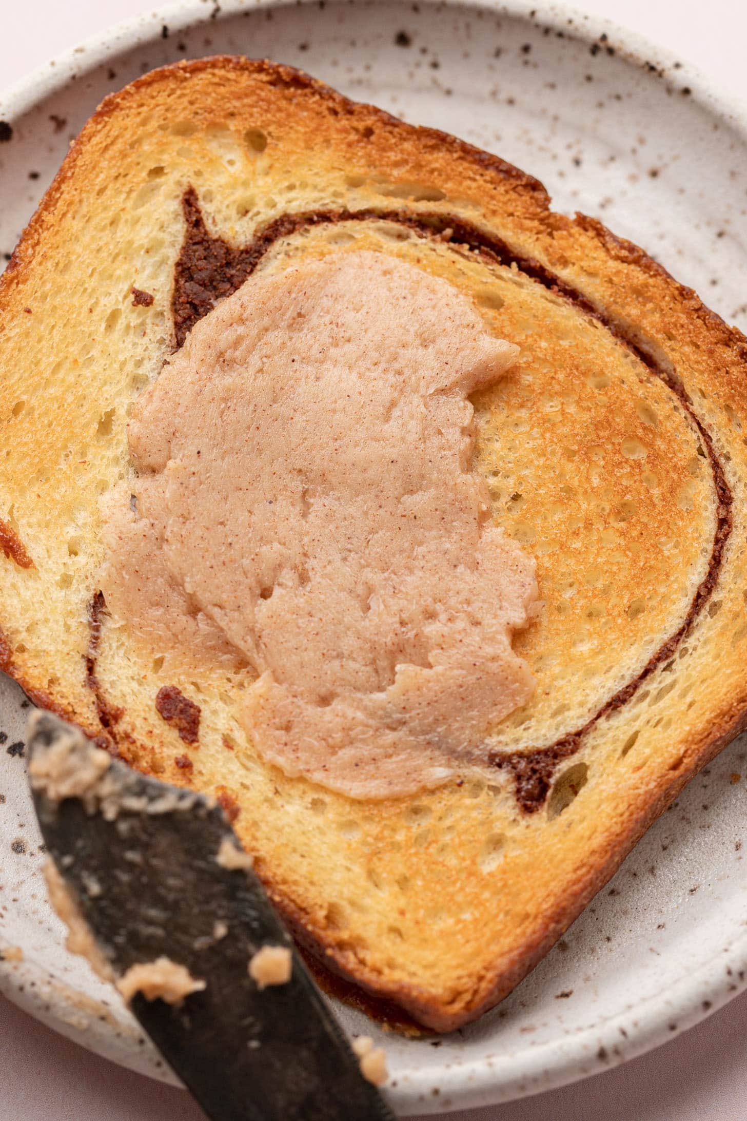 A slice of toasted bread with cinnamon spread on a plate, and a knife with more spread nearby.