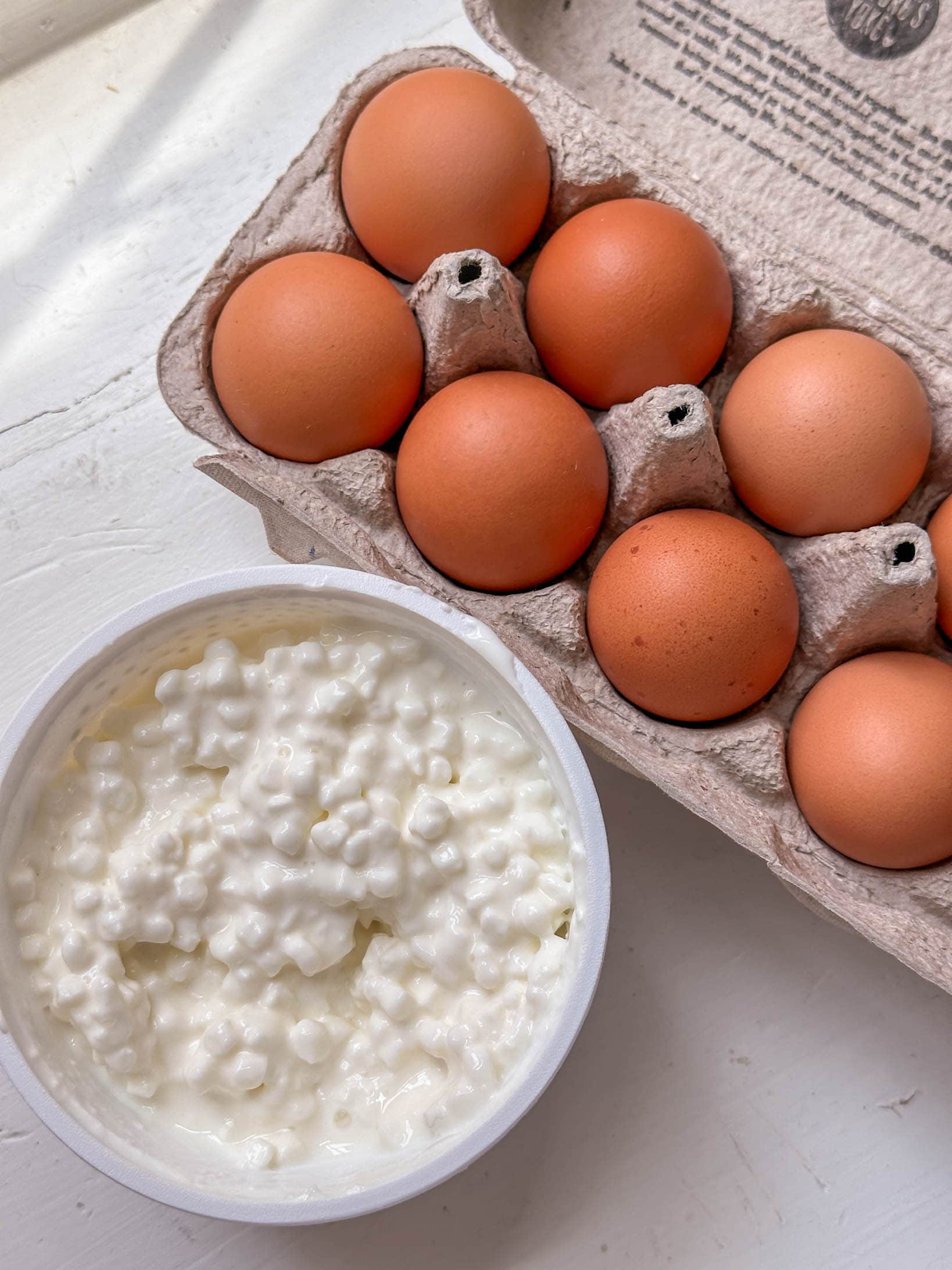 A carton of brown eggs and a bowl of cottage cheese on a white surface.