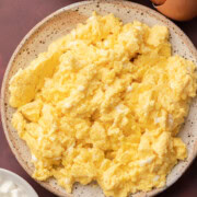 Plate of scrambled eggs with a bowl of eggs and some cottage cheese on a dark surface.