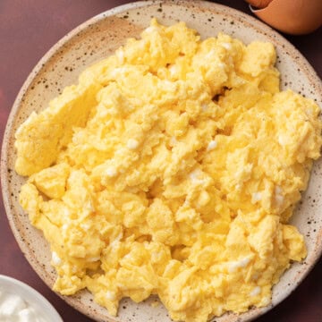 Plate of scrambled eggs with a bowl of eggs and some cottage cheese on a dark surface.