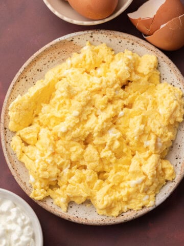 Plate of scrambled eggs with a bowl of eggs and some cottage cheese on a dark surface.