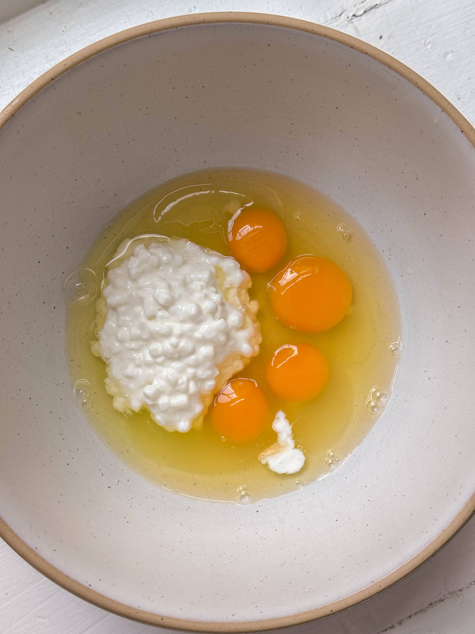 Four raw eggs and a scoop of cottage cheese in a beige bowl on a white surface.