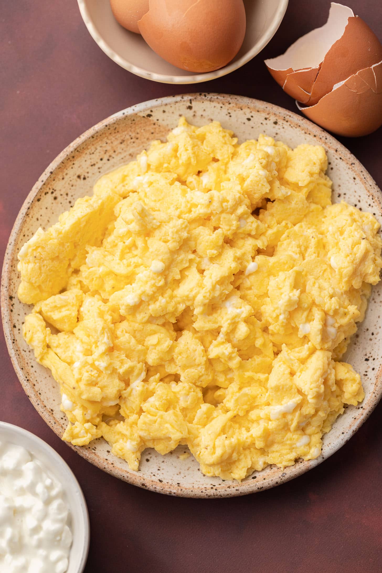 Plate of scrambled eggs with a bowl of eggs and some cottage cheese on a dark surface.