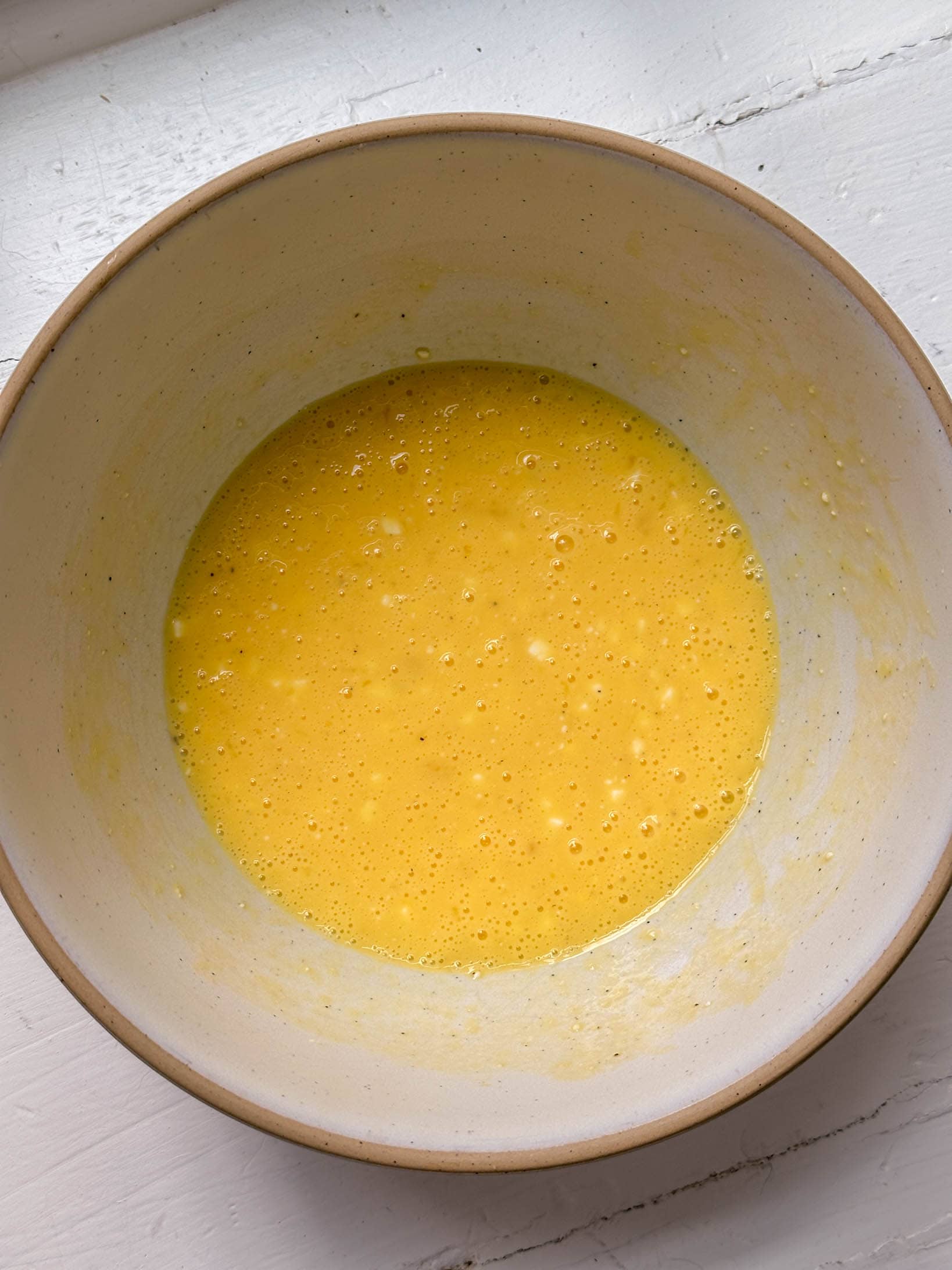 A white bowl with a yellow, whisked egg mixture on a white wooden surface.