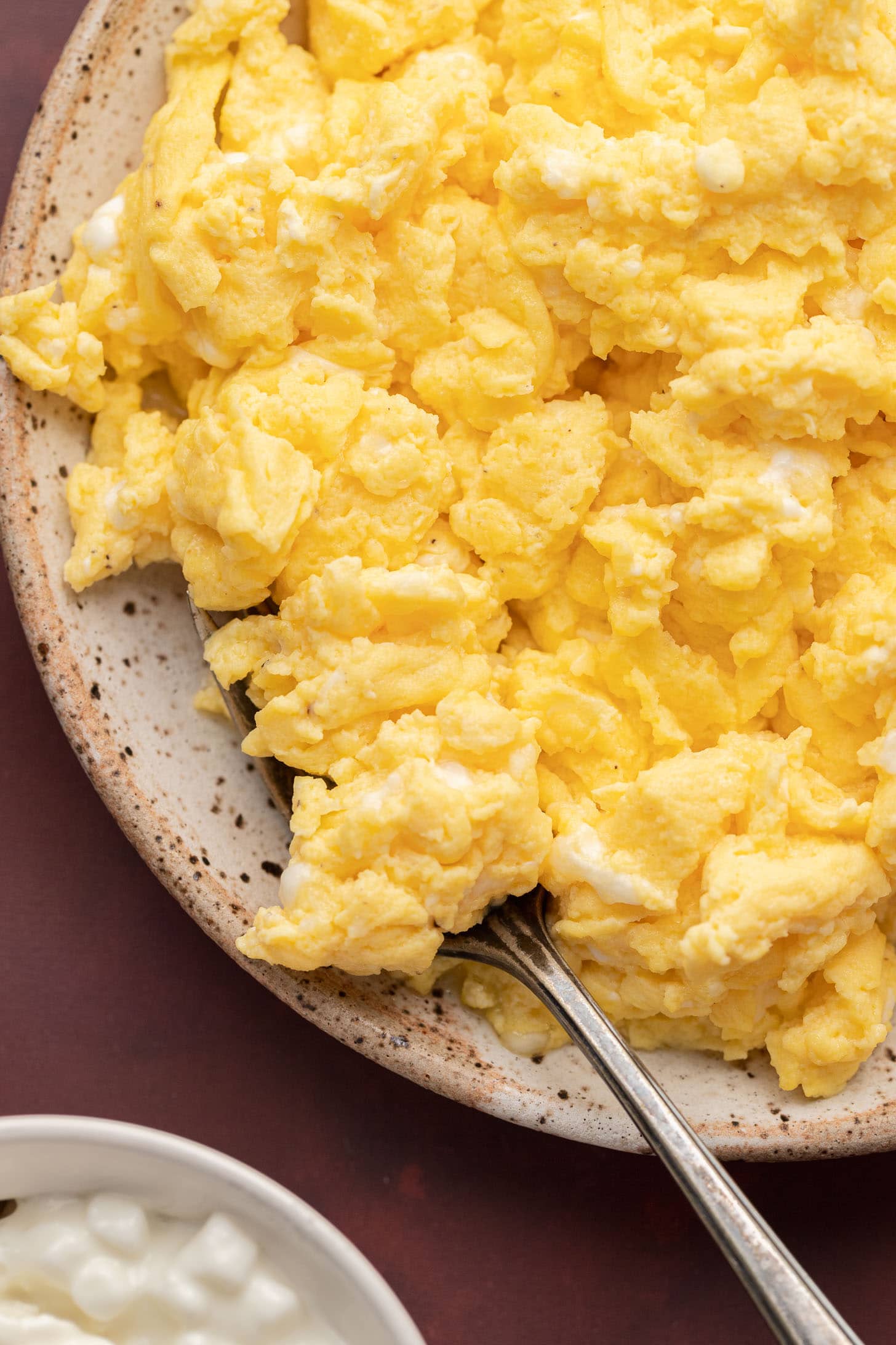 A plate of scrambled eggs with a spoon on a speckled ceramic dish, next to a bowl of cottage cheese.