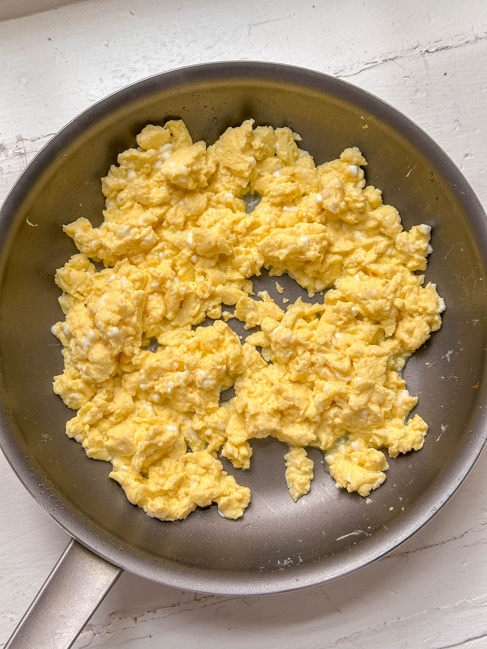 Scrambled eggs cooking in a nonstick frying pan on a white wooden surface.