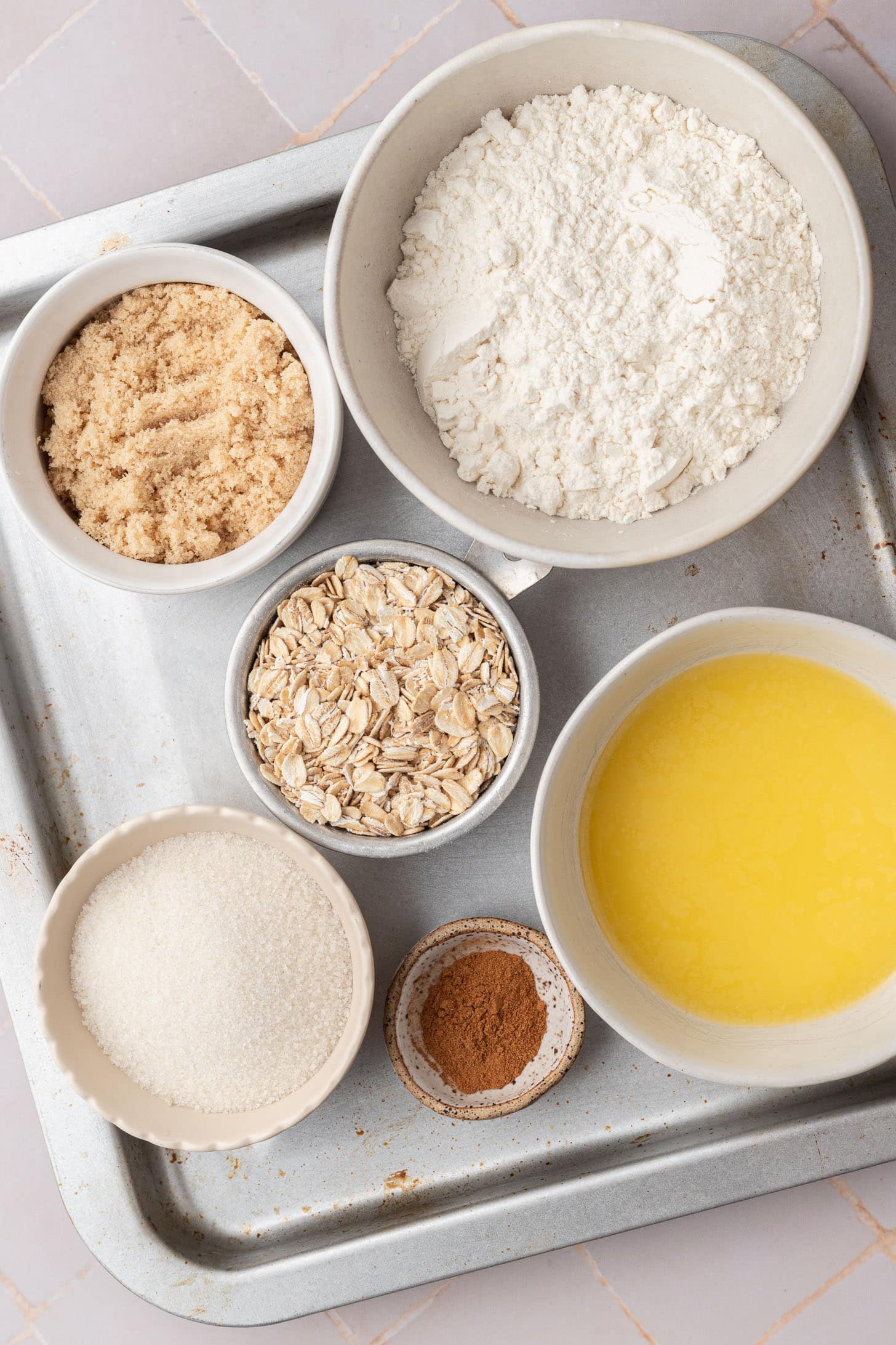 Five bowls with flour, brown sugar, oats, white sugar, melted butter, and a pinch of cinnamon on a tray.