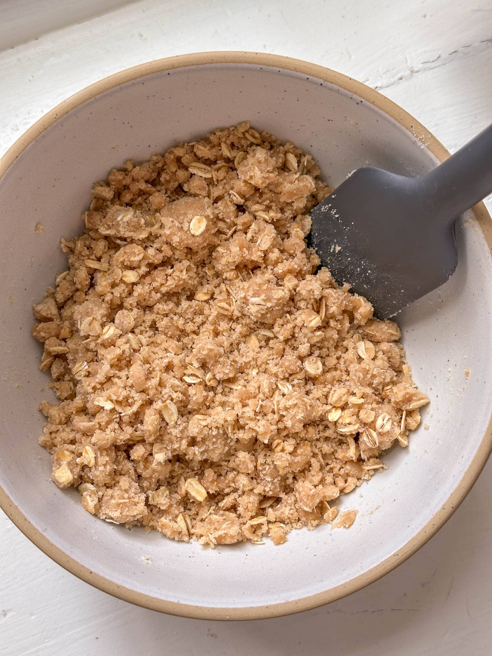 A gray spatula rests in a bowl filled with crumbly oat and brown sugar mixture on a white surface.